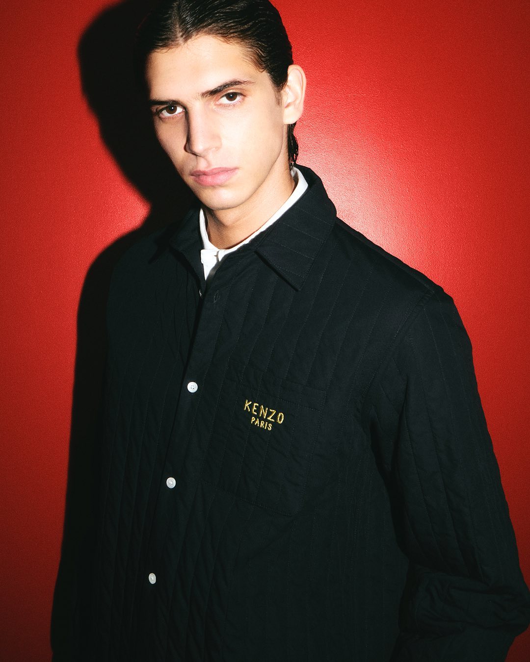 Close-up portrait of a male model wearing a black buttoned shirt with gold embroidered text against a red background.