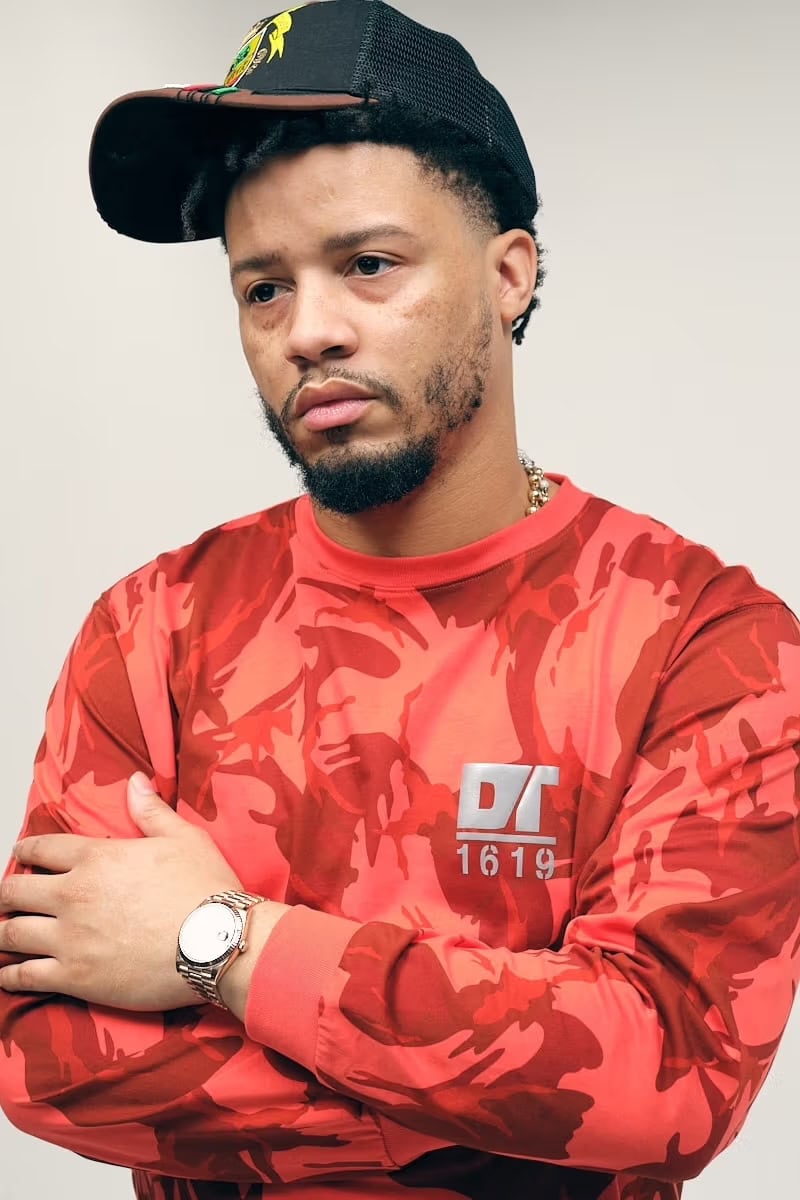 Veeze wearing a red camo sweatshirt and black cap with arms crossed against a white background for Denim Tears.