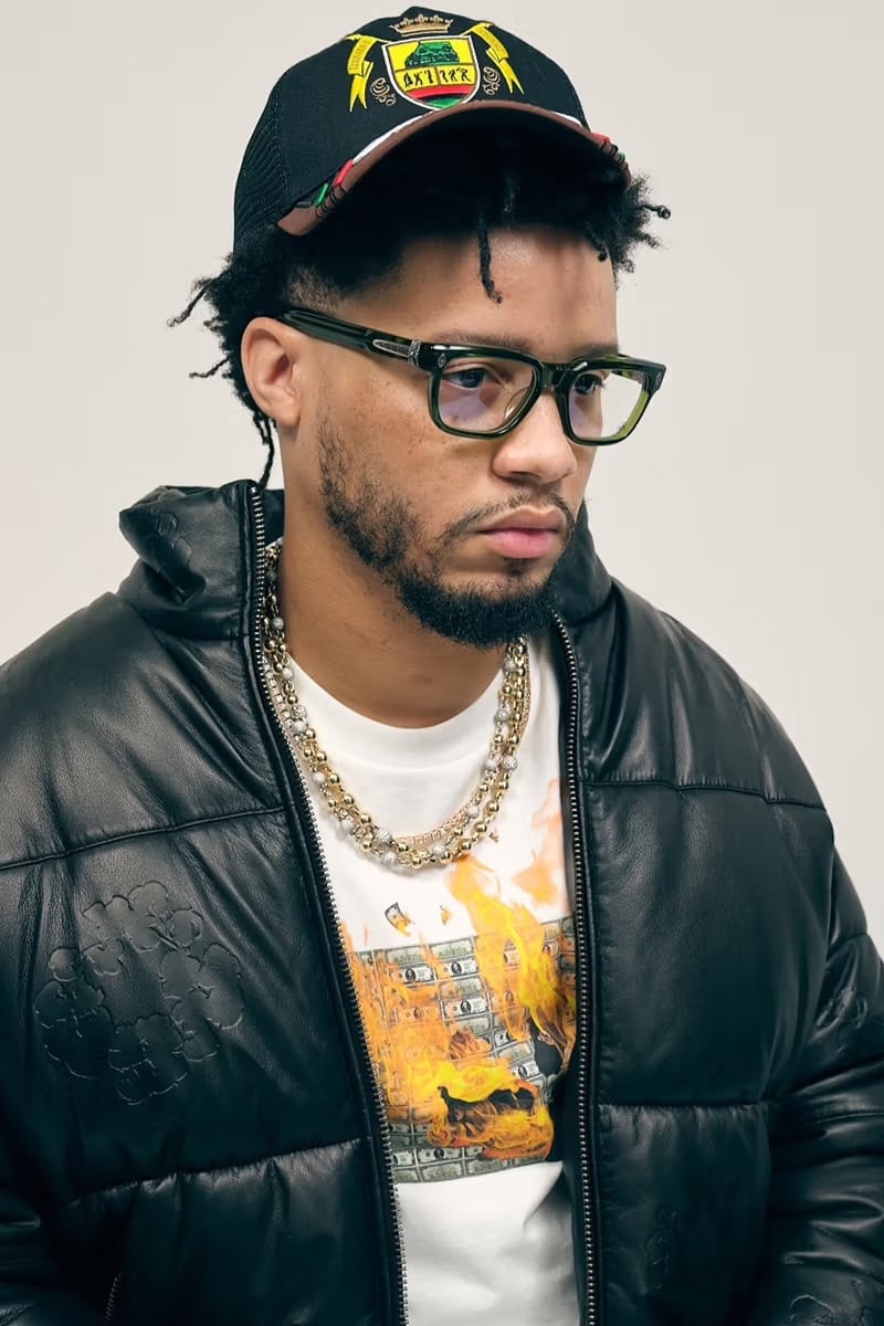 Man wearing a black puffer jacket, graphic t-shirt, layered necklaces, eyeglasses, and a baseball cap against a white background