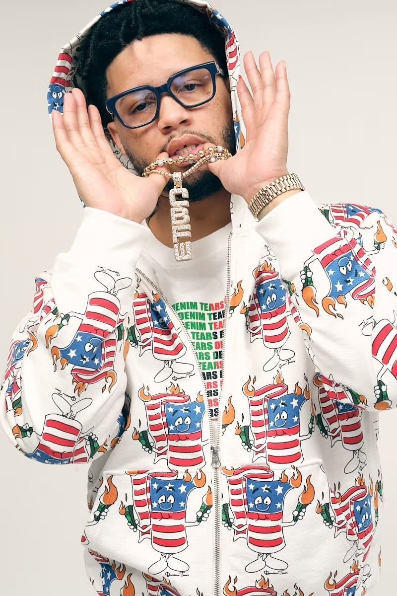 Man wearing patterned zip hoodie holding gold pendant necklace against plain white background