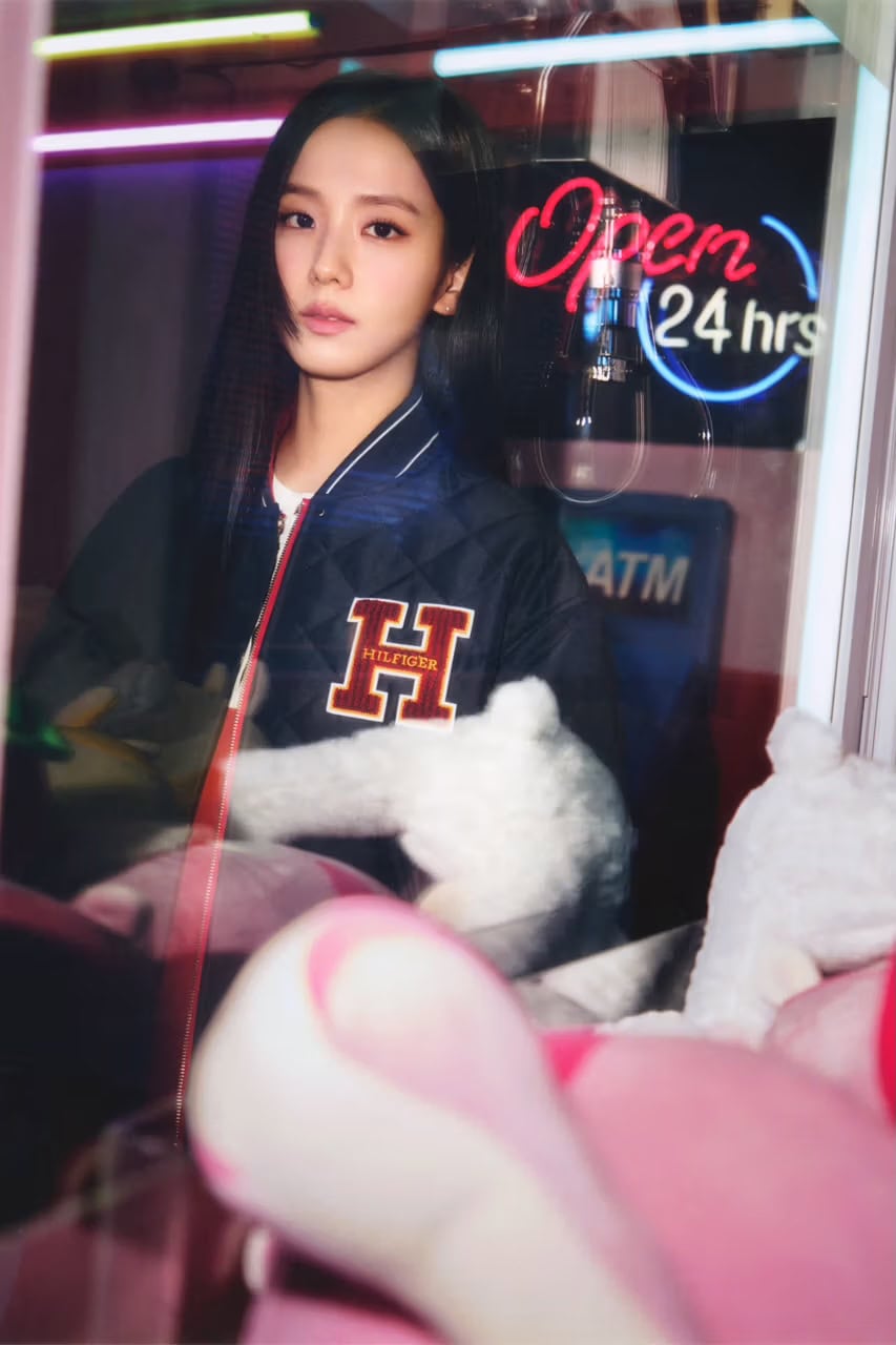 Jisoo wearing a varsity jacket featuring a prominent letter H patch behind the glass window of a claw machine.