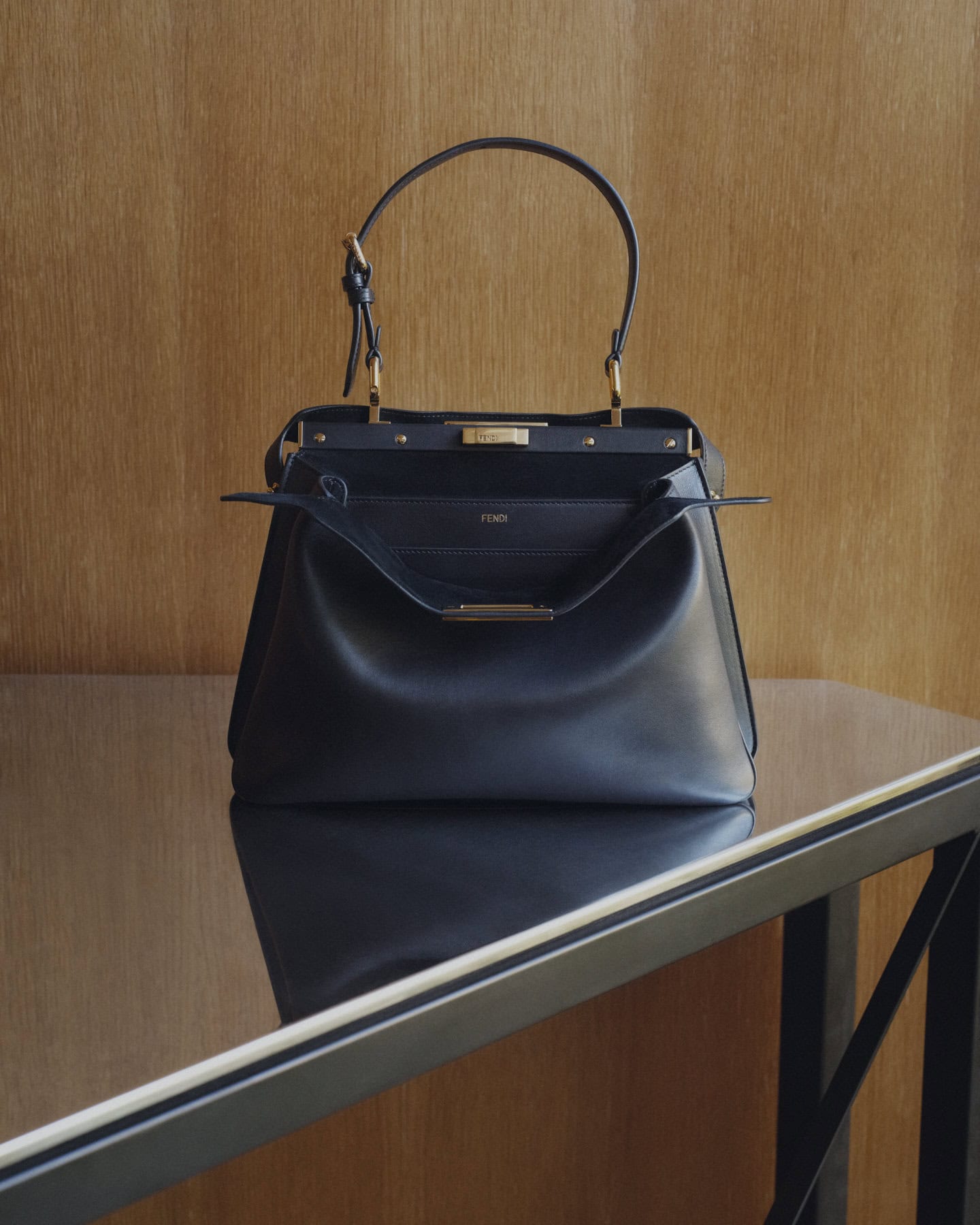 Black Fendi Peekaboo Soft Small leather handbag resting on a reflective dark tabletop against a warm wood paneled wall.