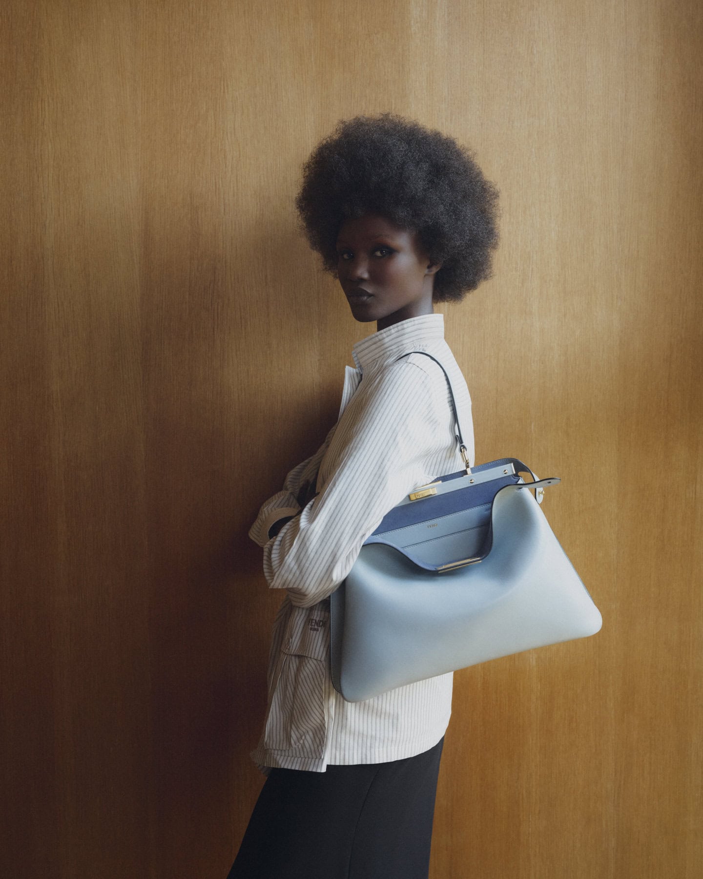 Model with afro hair wearing a striped shirt and carrying a blue Fendi Peekaboo Soft Small Bag against wood paneling