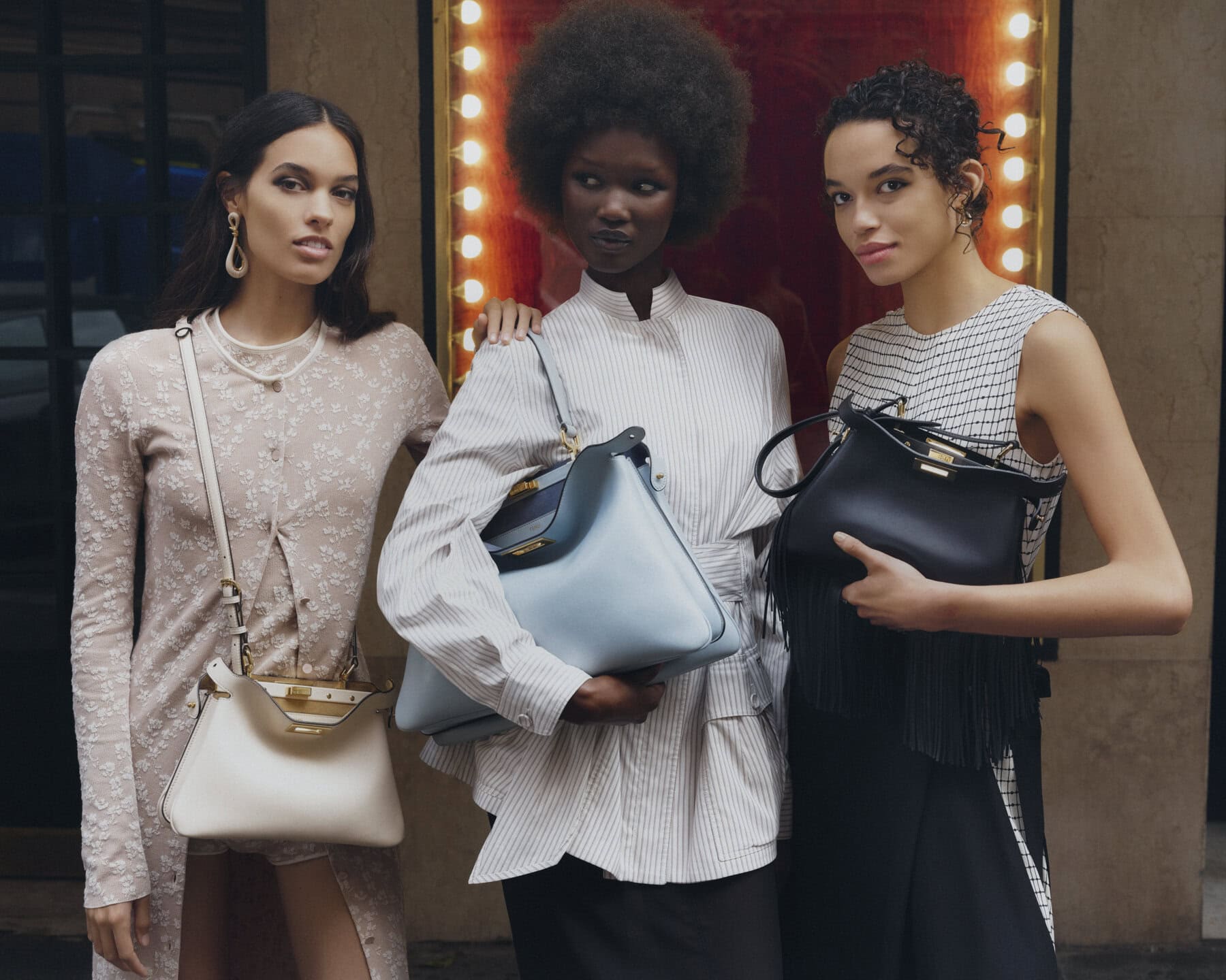 Three female models posing together outdoors while holding Fendi Peekaboo handbags in white, light blue, and black.