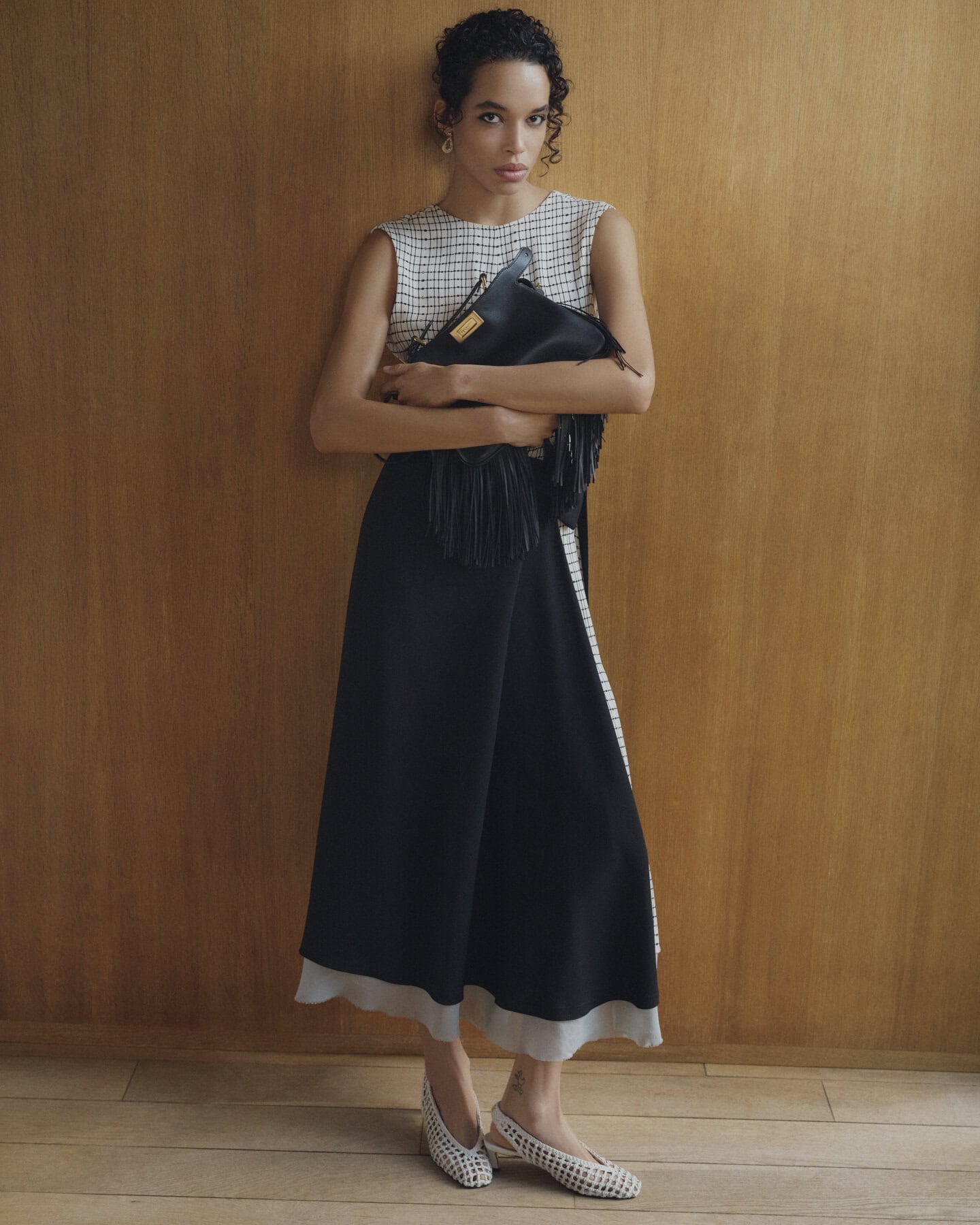 Model standing against wood paneling holding a black fringed handbag, wearing a patterned top and black skirt.