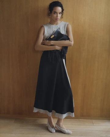 Model standing against wood paneling holding a black fringed handbag, wearing a patterned top and black skirt.