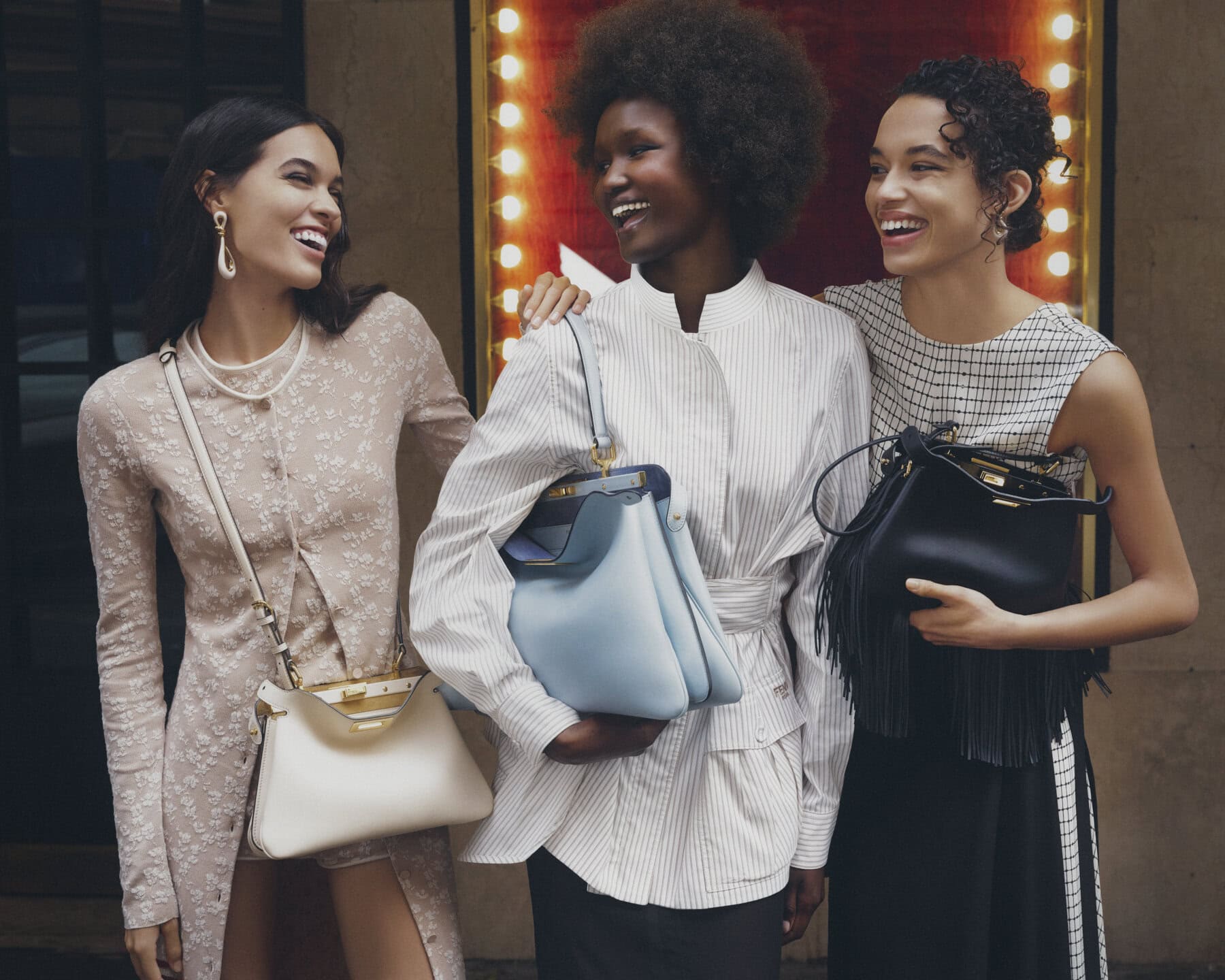 Three smiling models posing together outdoors, holding Fendi Peekaboo Soft handbags against a building entrance with warm lights.