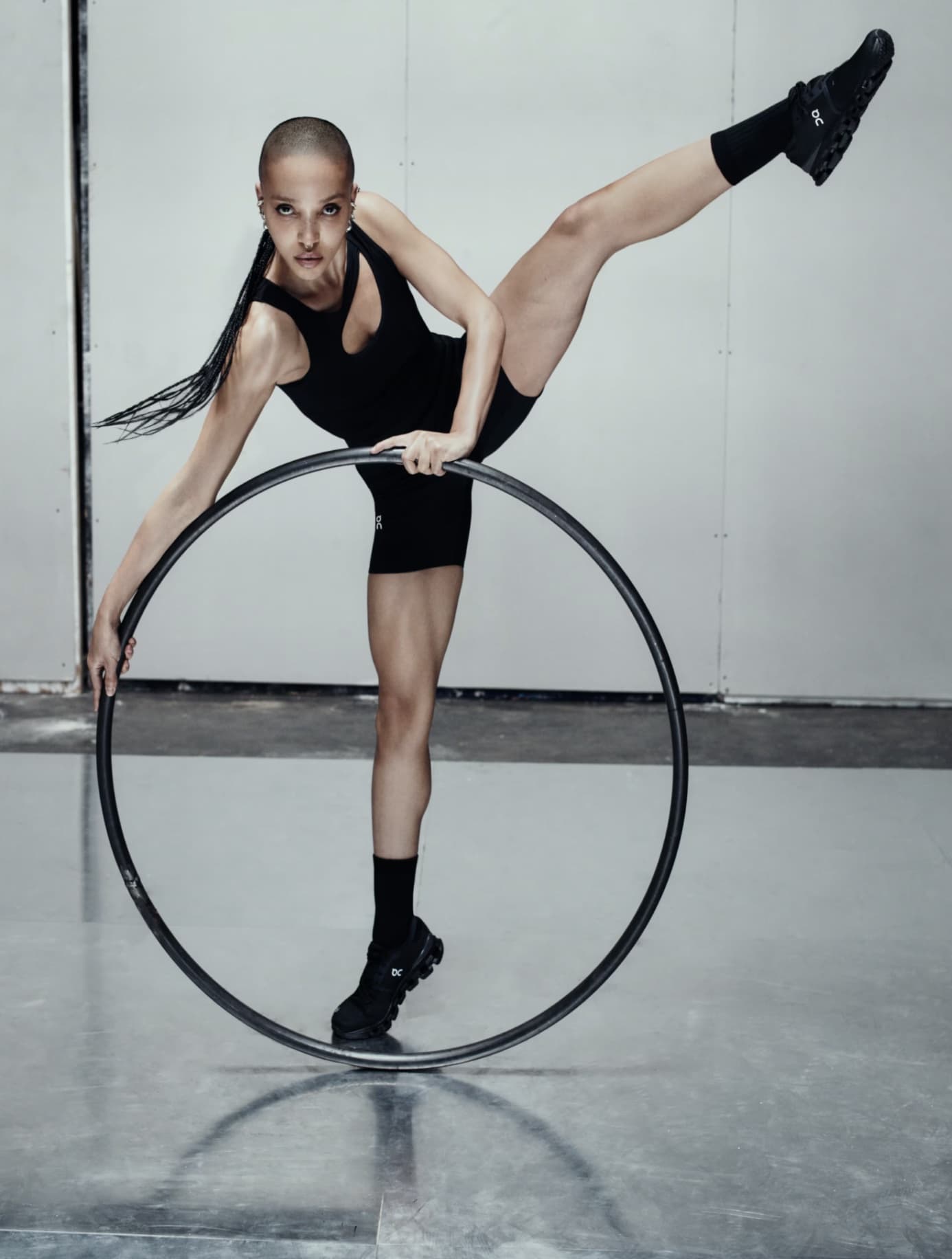 FKA Twigs posing with a hula hoop wearing a black athletic outfit and sneakers against a concrete background