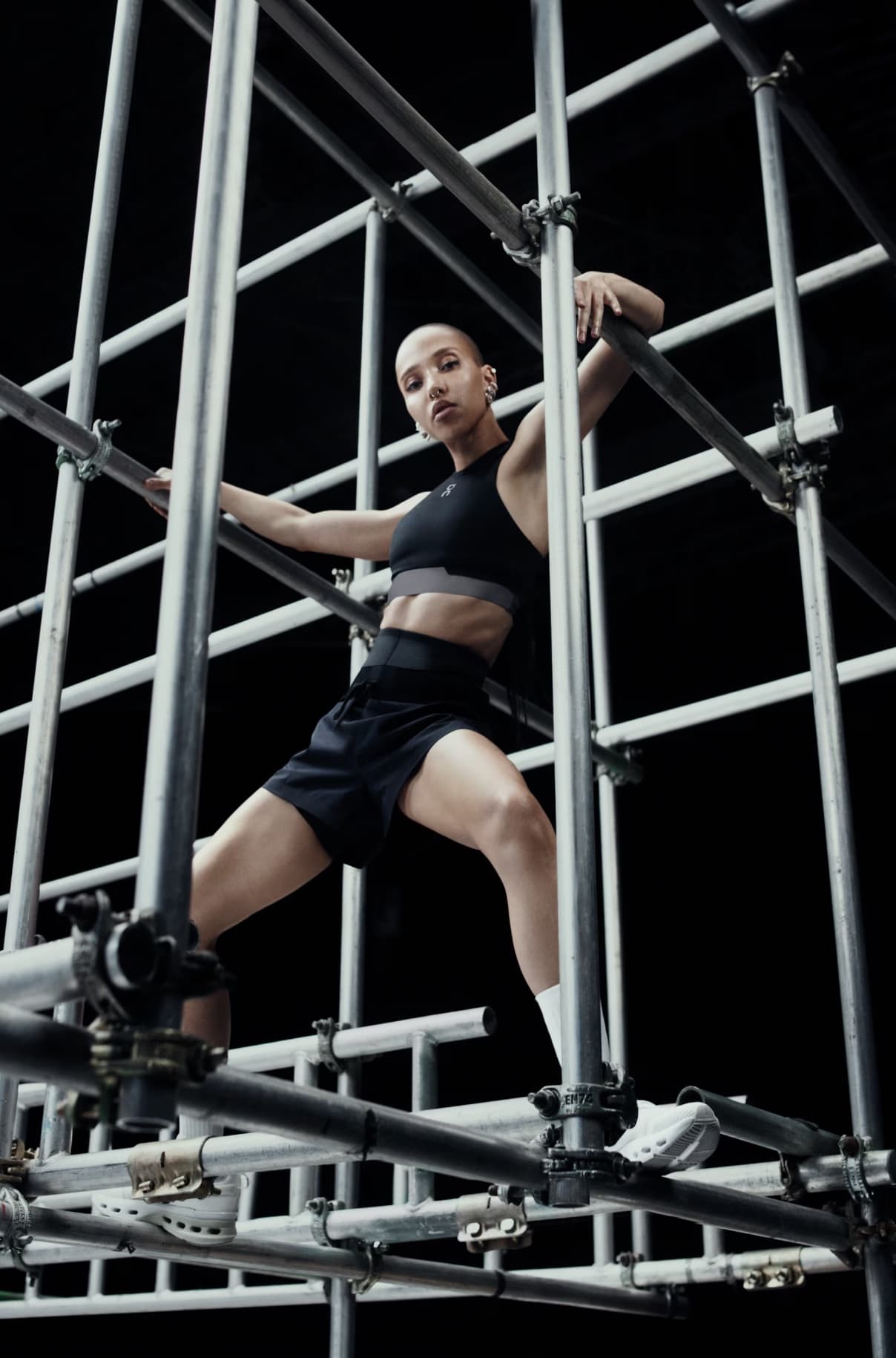 Model posing inside a metal scaffolding structure wearing black athletic crop top and shorts against a dark background