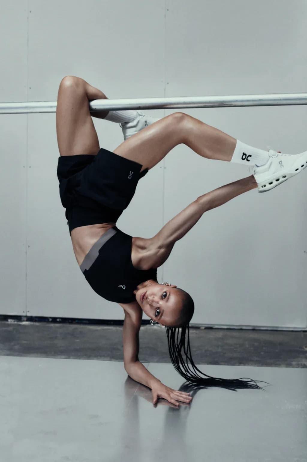 Model doing a handstand holding onto a horizontal bar wearing black athletic wear and white sneakers against a neutral grey studio background