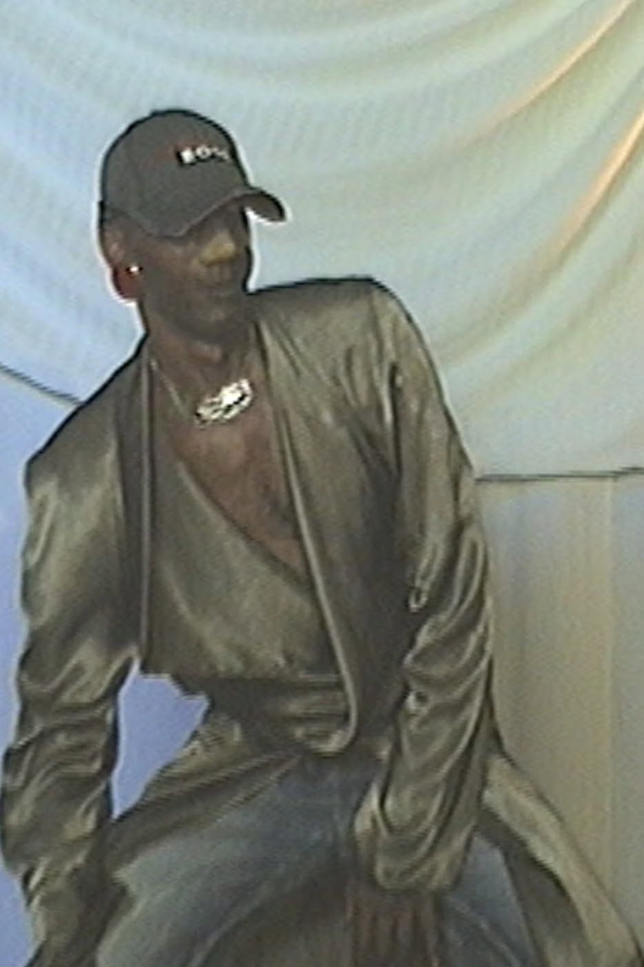 Man wearing a baseball cap, silver necklace, and metallic gold draped jacket against a textured white backdrop.