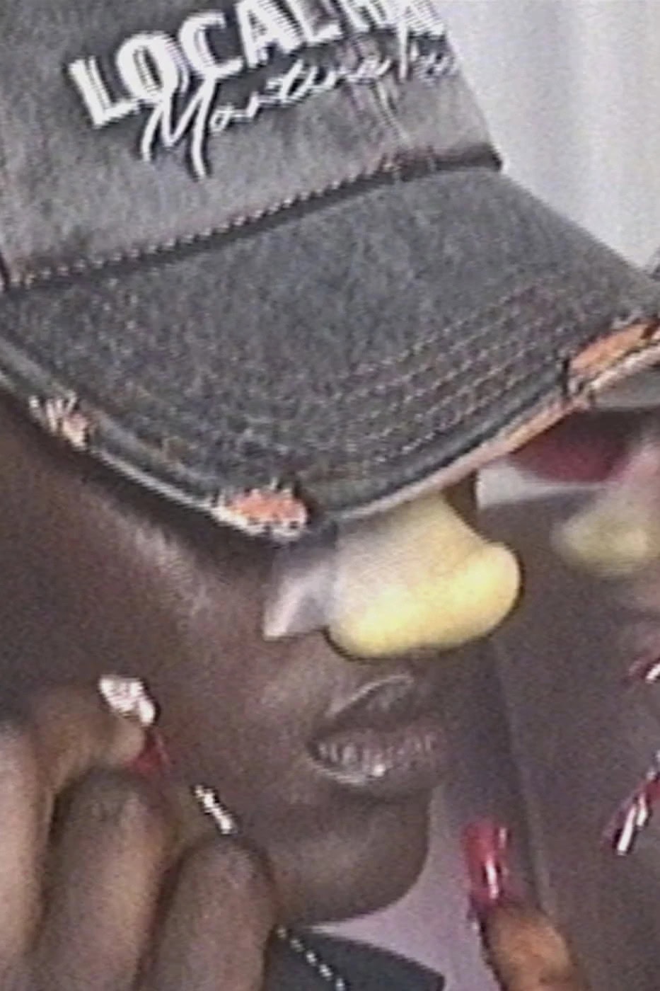 Close-up portrait of a person wearing a dark baseball cap and a prosthetic nose, photographed by Sharna Osborne for Martine Rose.
