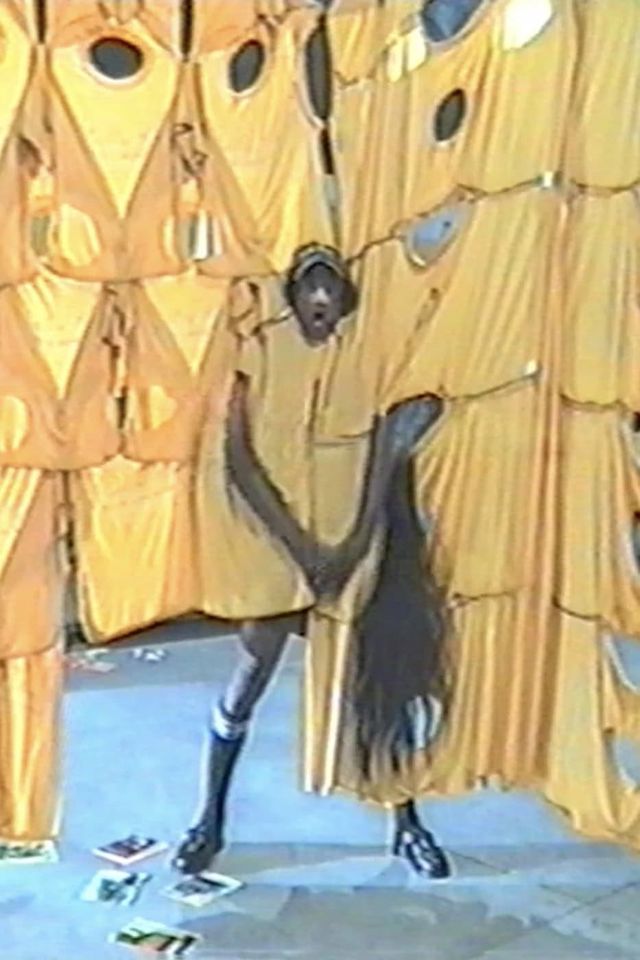 A person standing in front of draped yellow fabric panels wearing dark garments and tall boots
