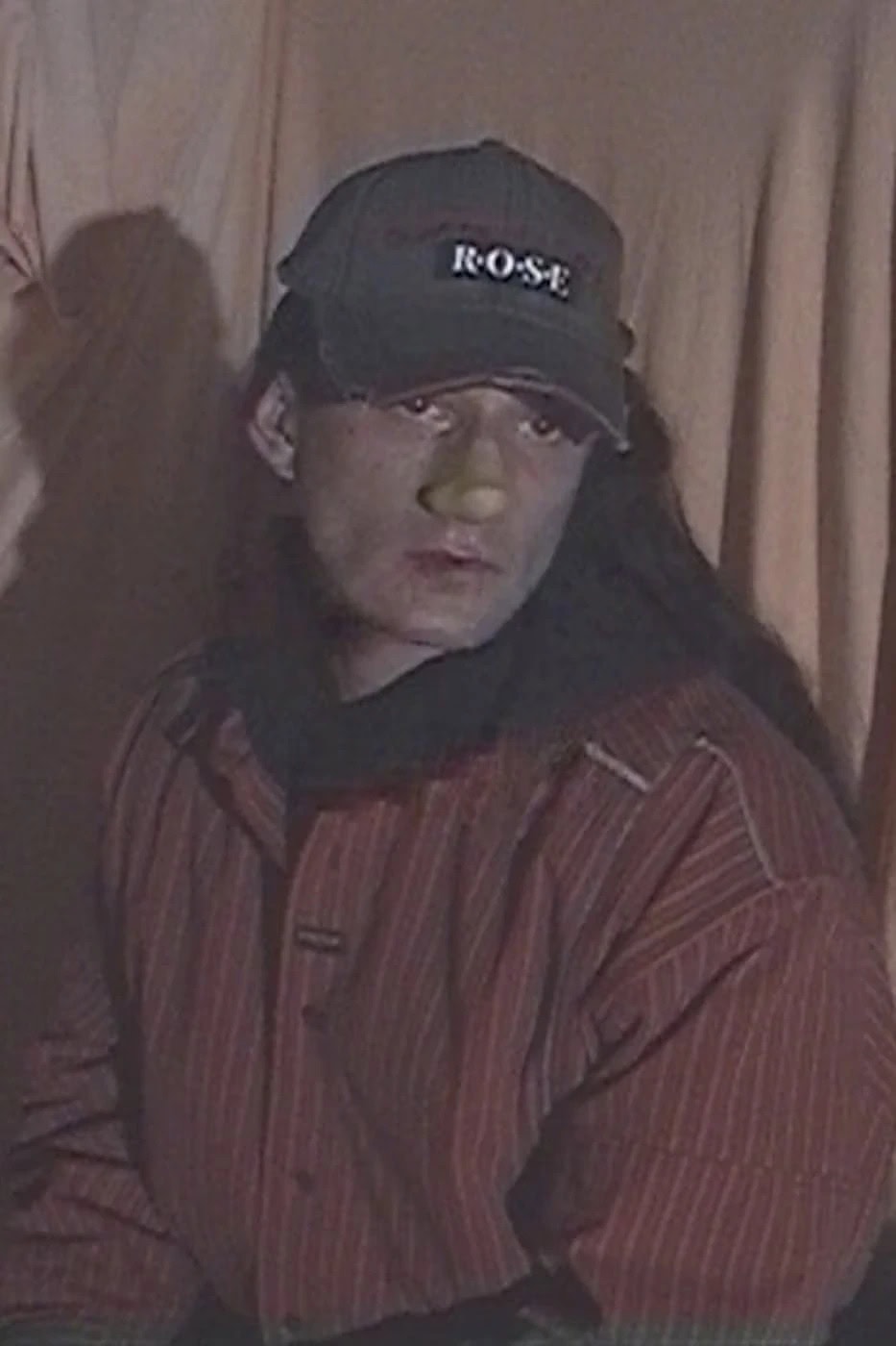Man with long dark hair wearing a striped red shirt and a dark cap featuring the word ROSE against a draped curtain background