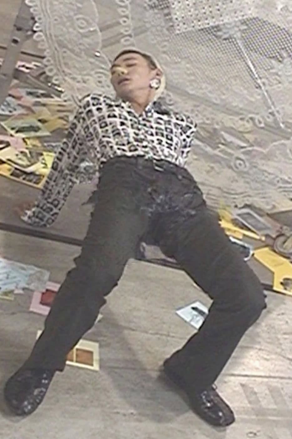 Model wearing patterned shirt and dark trousers leaning back against a set surface with papers scattered around.