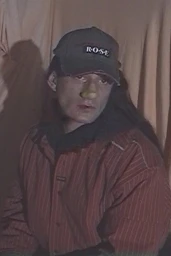 Man with long dark hair wearing a striped red shirt and a dark cap featuring the word ROSE against a draped curtain background