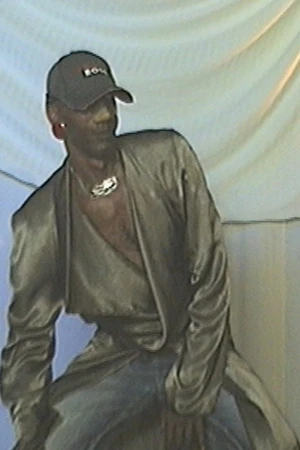 Man wearing a baseball cap, silver necklace, and metallic gold draped jacket against a textured white backdrop.