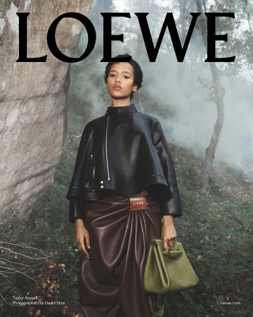 Model Taylor Russell in a black leather jacket holding an olive green handbag standing outdoors against rocky terrain.
