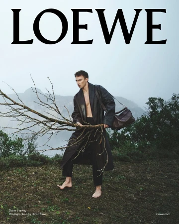 Drew Starkey holding a bare branch in a dark leather coat outdoors for Loewe