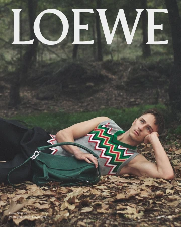 Drew Starkey lying on a forest floor of dry leaves wearing a patterned knit vest and dark trousers next to a green leather bag.