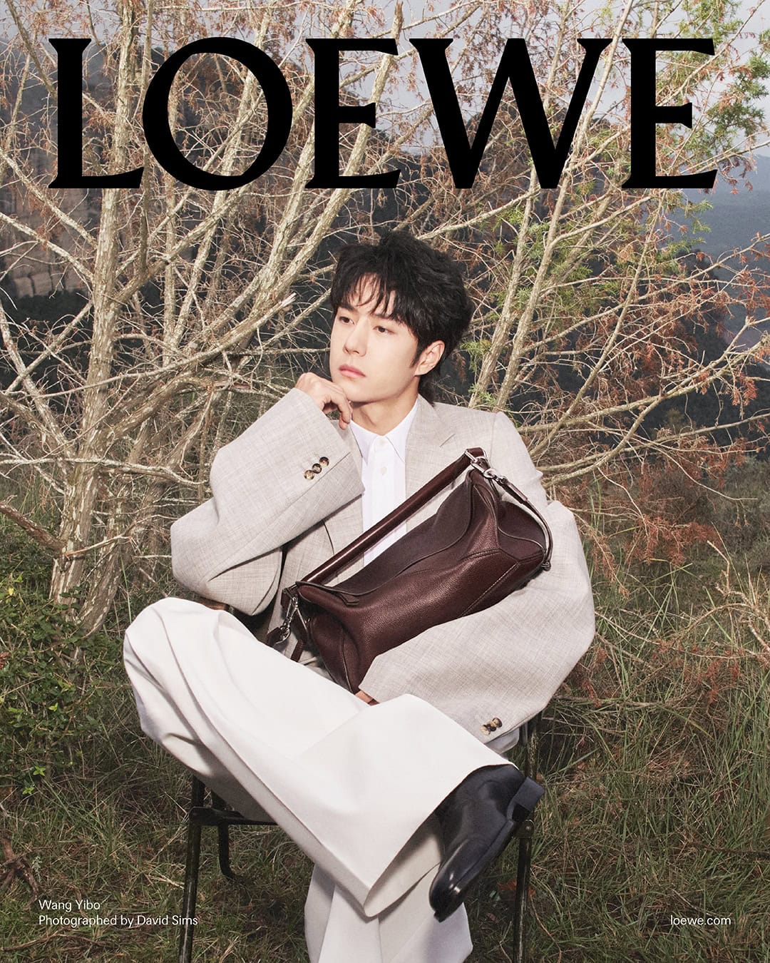 Wang Yibo seated outdoors wearing a light blazer and white trousers holding a brown leather bag for Loewe.
