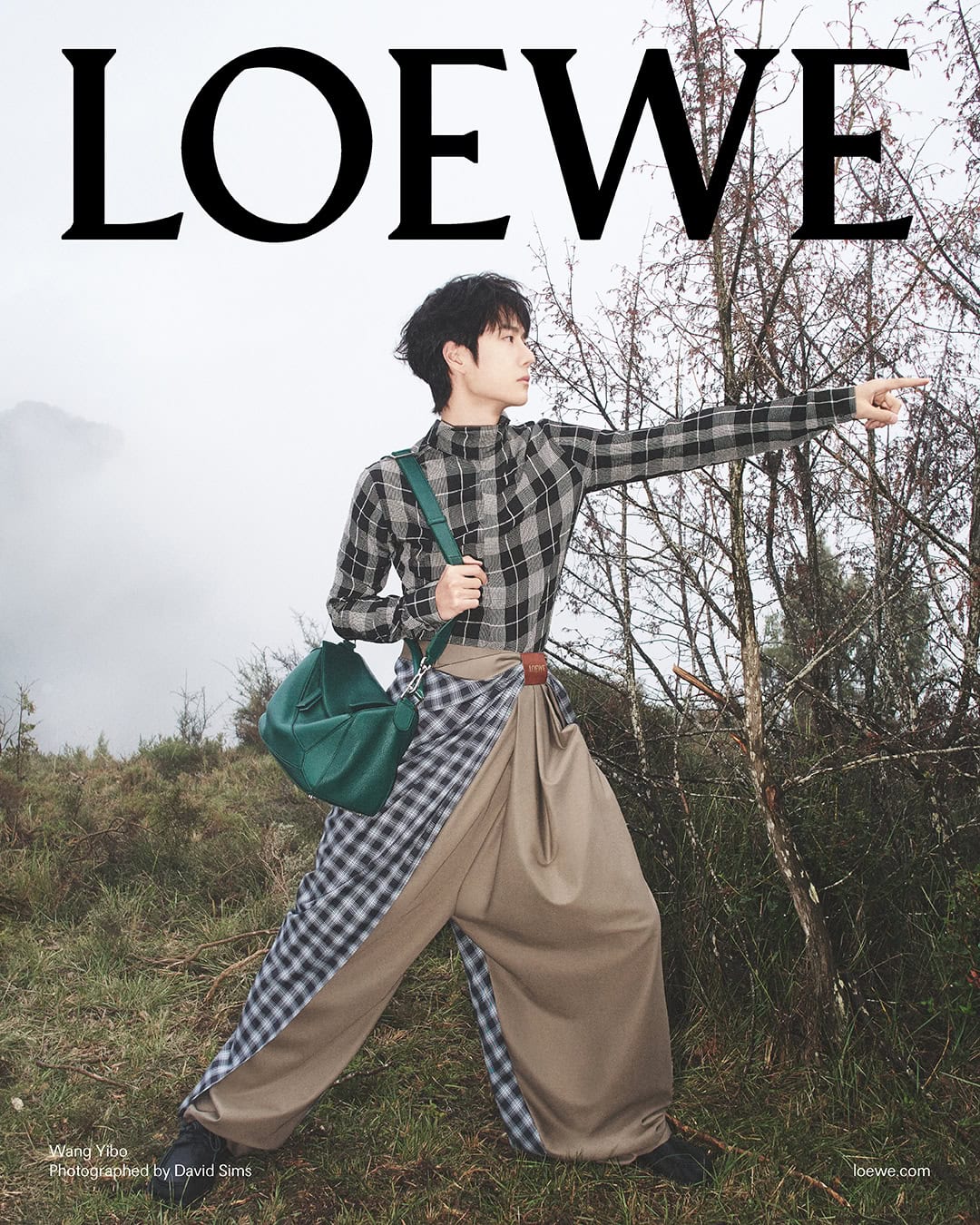 Wang Yibo in plaid shirt and draped trousers pointing outdoors carrying a green shoulder bag in an ad campaign by David Sims for Loewe.