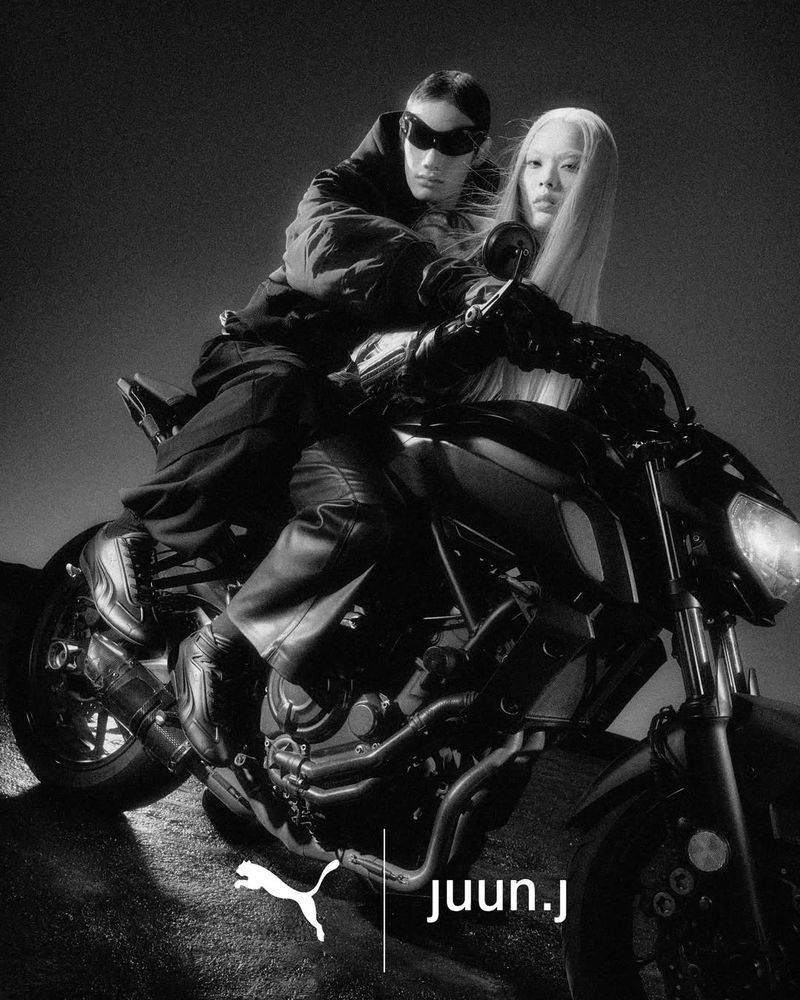 Two models posing on a black motorcycle in a grainy monochrome fashion campaign image with visible brand logos at the bottom.