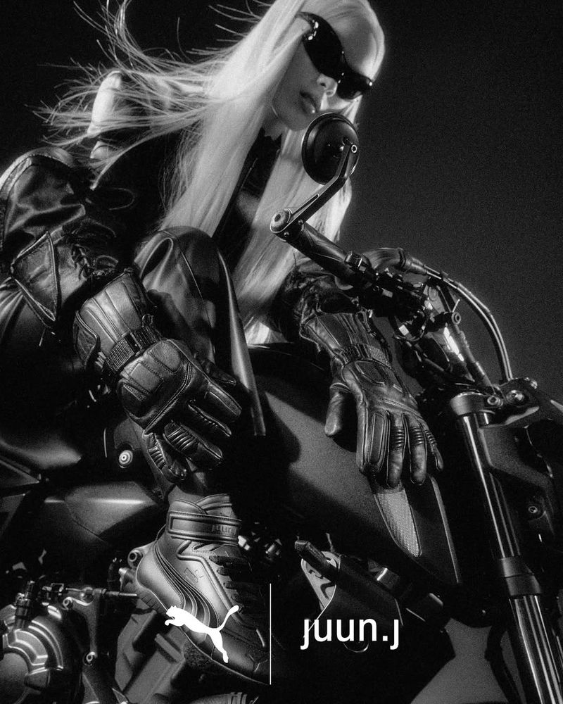 Monochrome fashion campaign image featuring a model in dark sunglasses and leather apparel seated on a motorcycle.