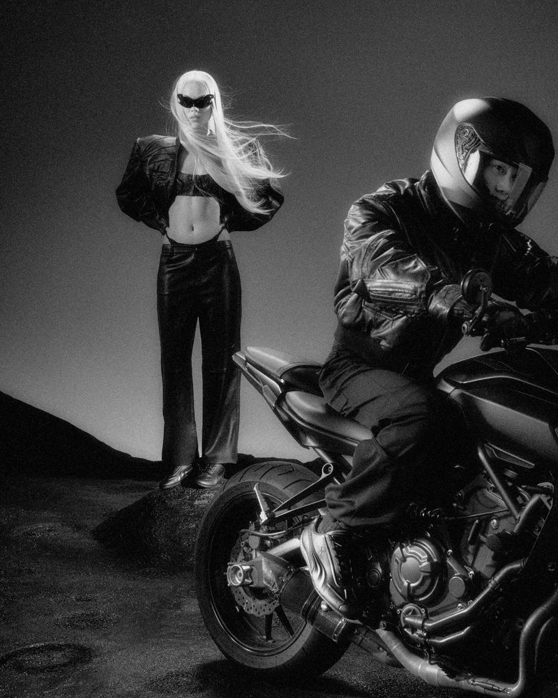 Monochrome fashion campaign image featuring a blonde woman standing behind a motorcyclist on a motorcycle in a studio set.