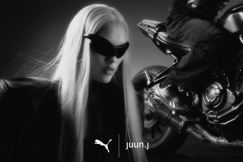 Model with long blonde hair wearing wraparound sunglasses and dark jacket beside a motorcycle for Juun.J x Puma Speedcat campaign