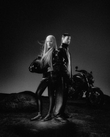 Two models wearing dark leather apparel posing next to a motorcycle under dramatic lighting in an outdoor setting