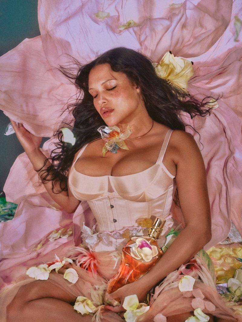 Model Alva Claire cradles a Jean Paul Gaultier perfume bottle amidst soft pink floral petals and draped fabric.