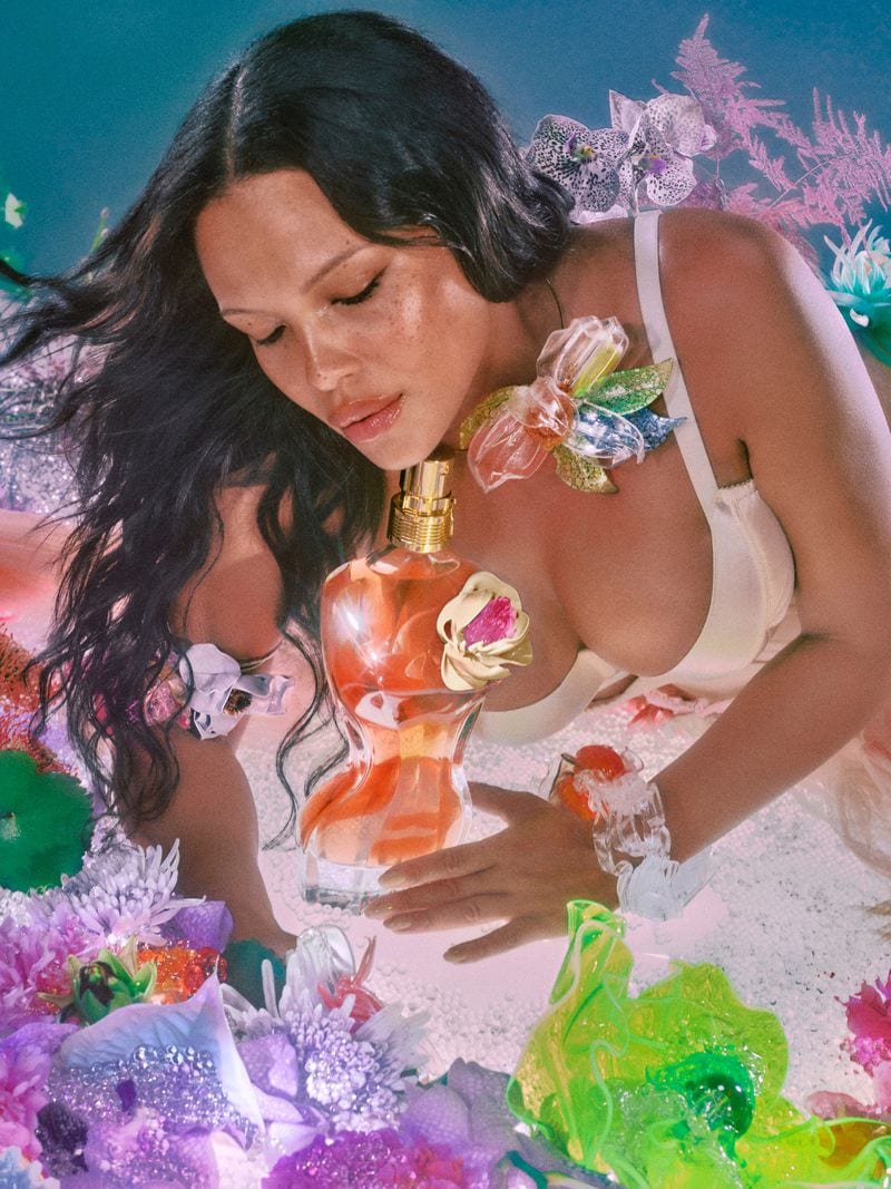 Model holding a torso-shaped perfume bottle amidst colorful floral displays in an ad campaign by Elizaveta Porodina for Jean Paul Gaultier