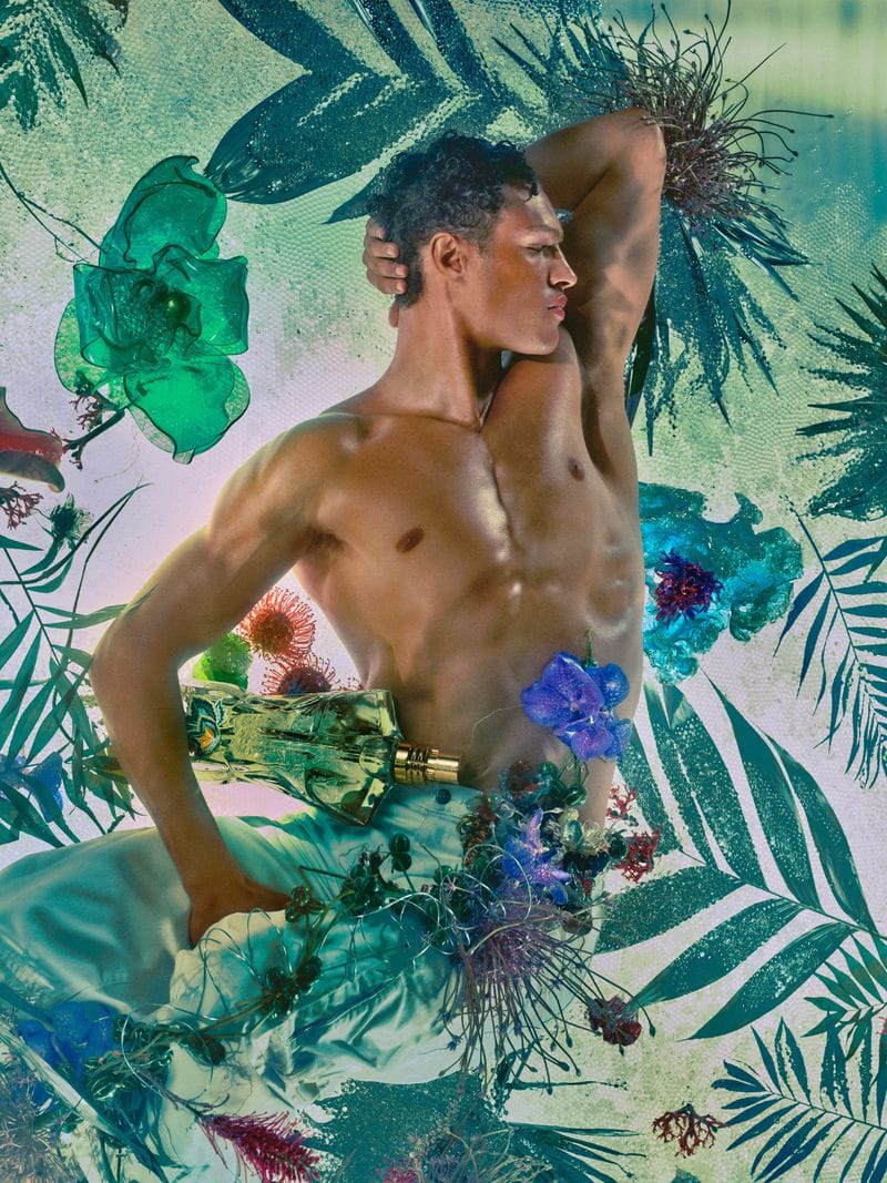 Shirtless male model posing amidst botanical green foliage and floral elements holding a perfume bottle for Jean Paul Gaultier