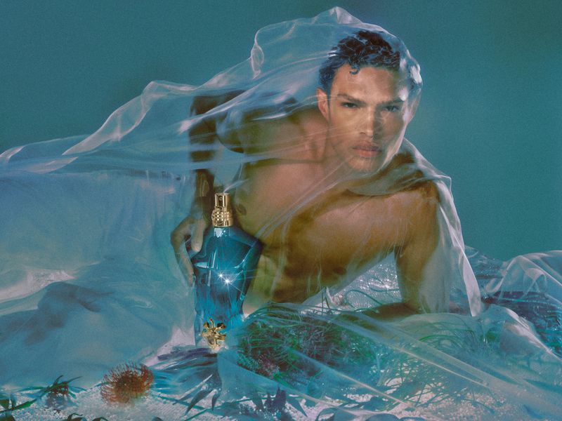 Shirtless man wrapped in translucent blue fabric holding a perfume bottle underwater in an editorial campaign image