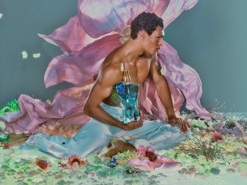 Shirtless male model holding a glass perfume bottle amidst a surreal floral set designed for Jean Paul Gaultier.