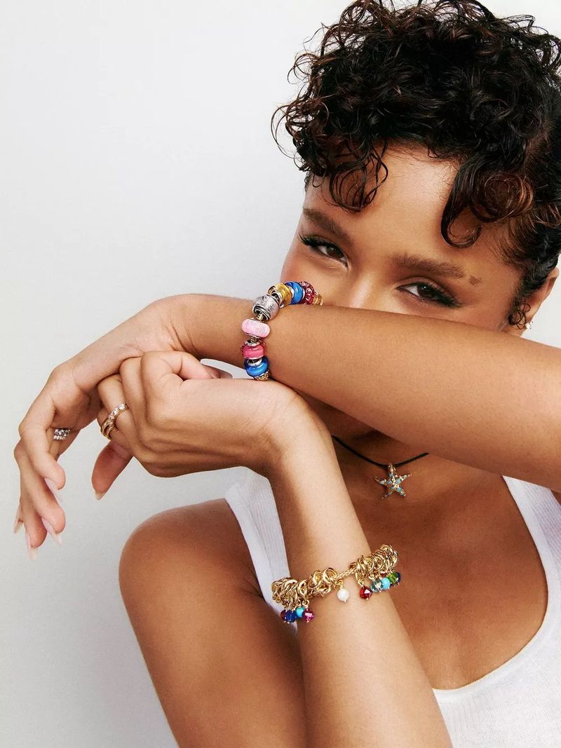 Portrait of a woman with curly hair in a white tank top, wearing two charm bracelets on each arm against a neutral background.