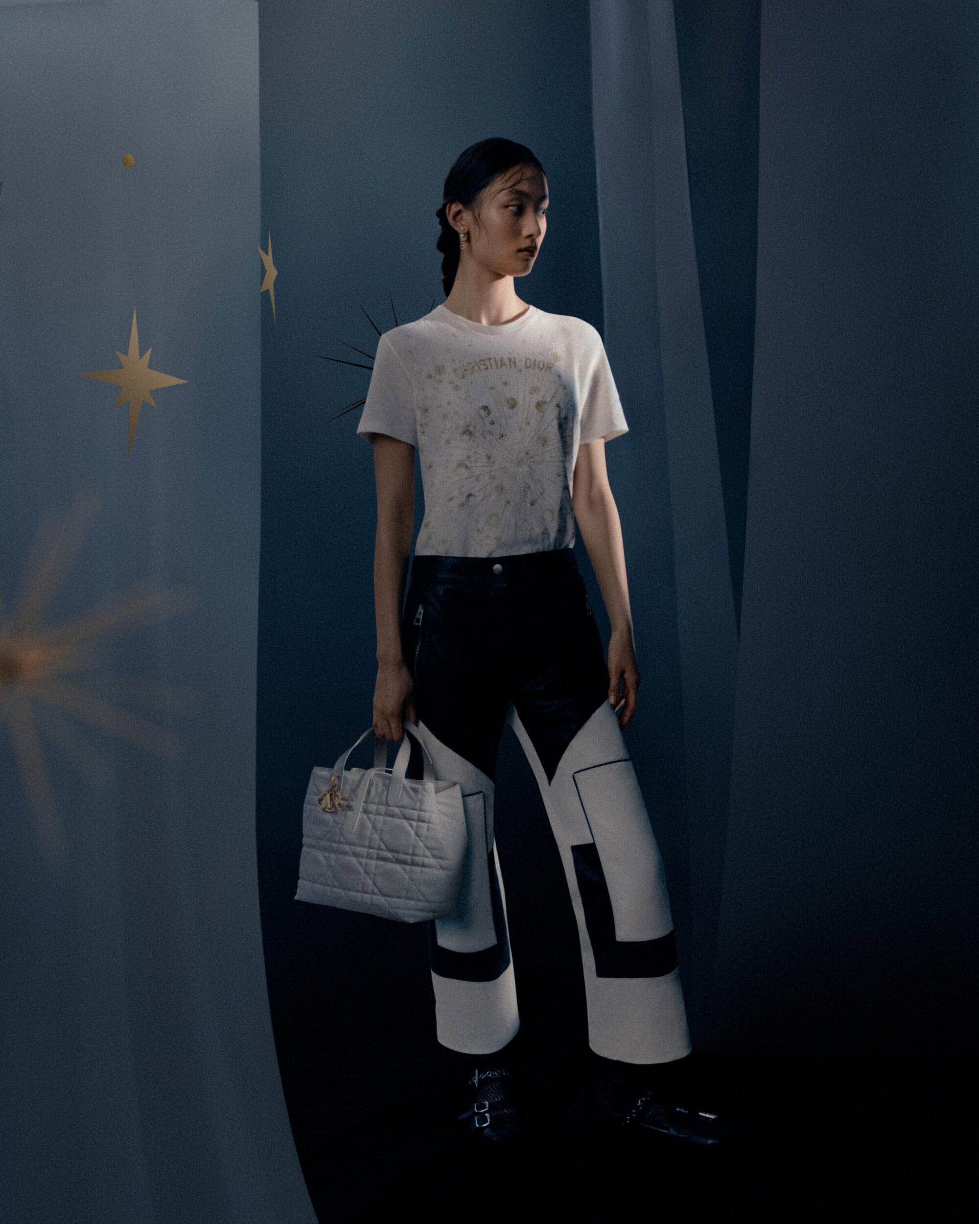 Model in graphic white t-shirt and patterned trousers holding a quilted tote bag against a moody background with gold stars