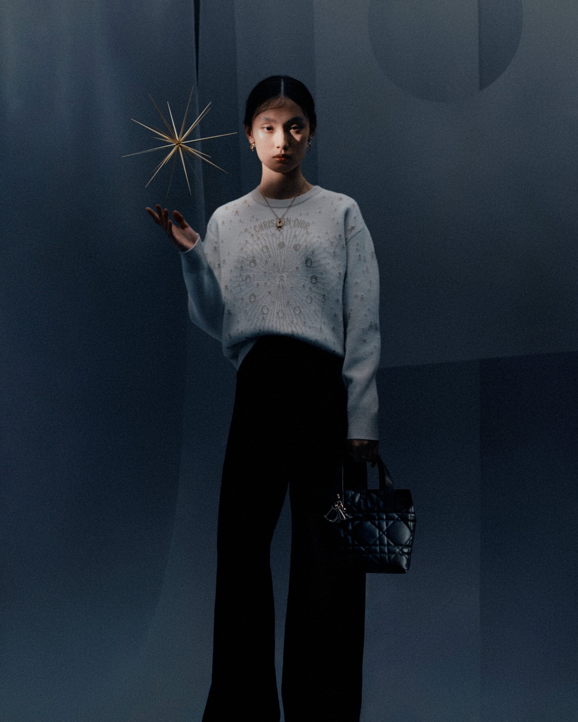 Model wearing a light grey sweater and black trousers holding a dark quilted handbag against a moody grey studio backdrop.