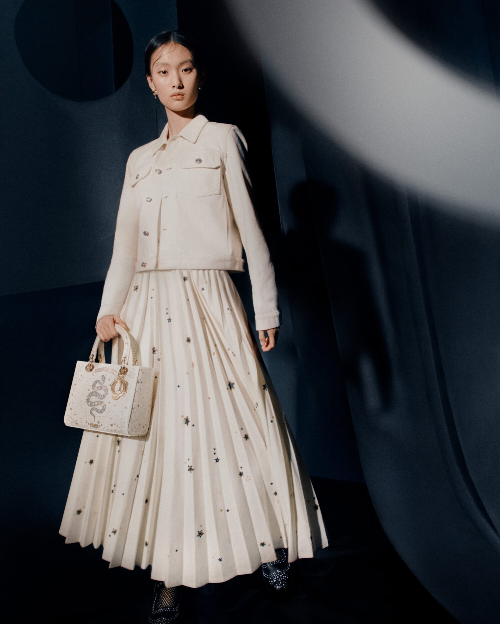 Model wearing a white pleated skirt and jacket holding an embroidered handbag against a dark backdrop