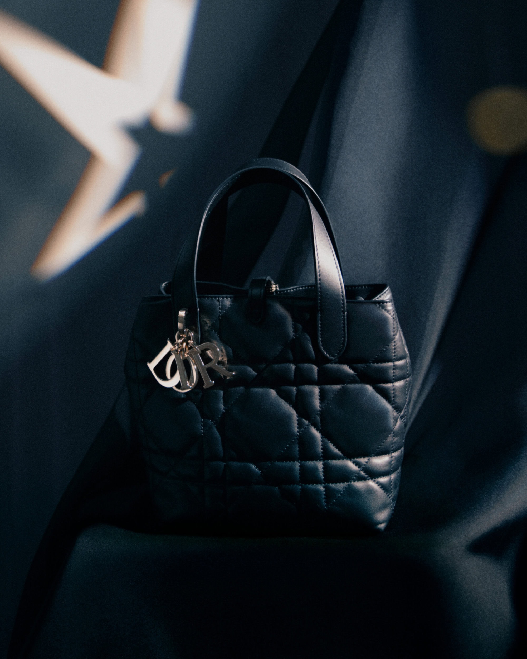Black quilted leather Dior handbag with hanging metal charms against dark draped fabric and a lit star graphic in the background.