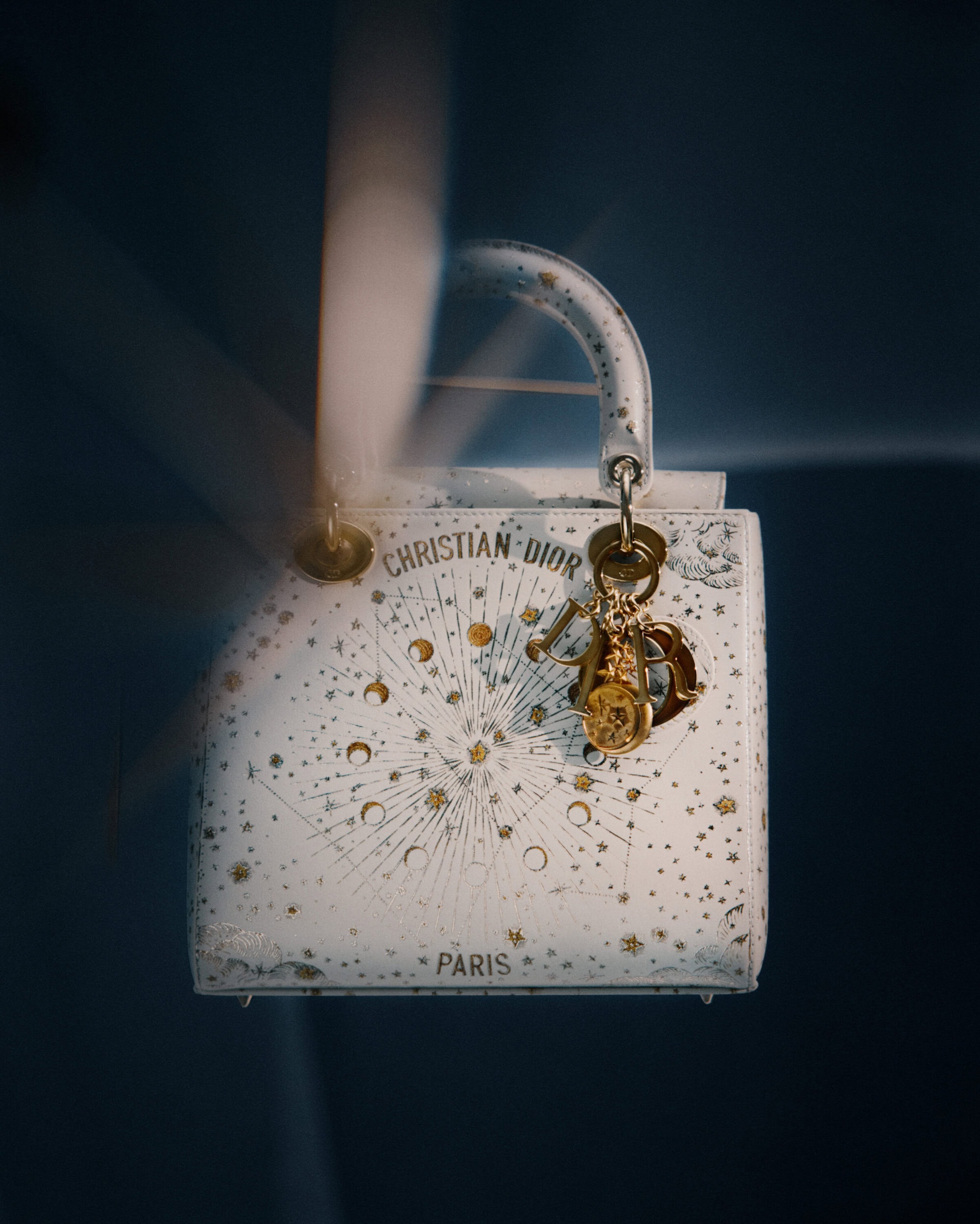 White leather Dior handbag with gold celestial motifs and charms against a dark gradient background.