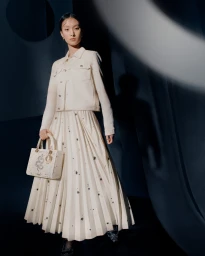 Model wearing a white pleated skirt and jacket holding an embroidered handbag against a dark backdrop