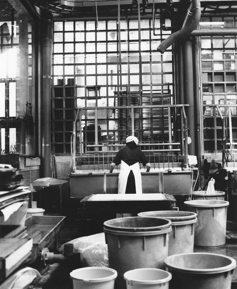 A person wearing a white apron works at a vat inside a factory setting with large tubs in the foreground.