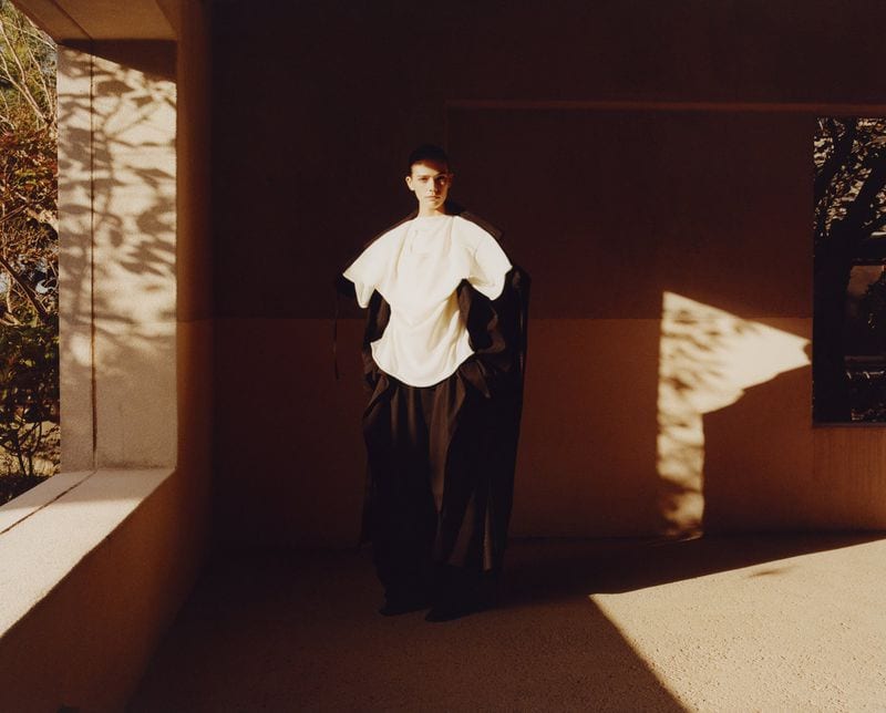 Model wearing a structured white top and dark coat standing in a shaded architectural alcove with sunlight casting dramatic patterns.