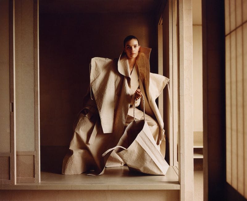 Model in voluminous sculptural beige coat holding a matching structured tote bag inside a traditional interior
