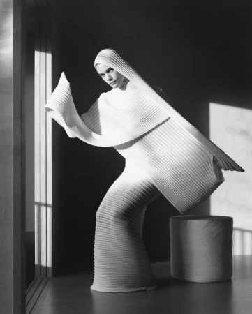 Model wearing a structured white pleated garment and matching hood posing in an interior space with strong directional light