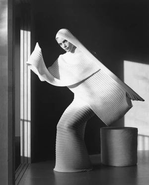 Model wearing a structured white pleated garment and matching hood posing in an interior space with strong directional light