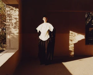 Model wearing a structured white top and dark coat standing in a shaded architectural alcove with sunlight casting dramatic patterns.