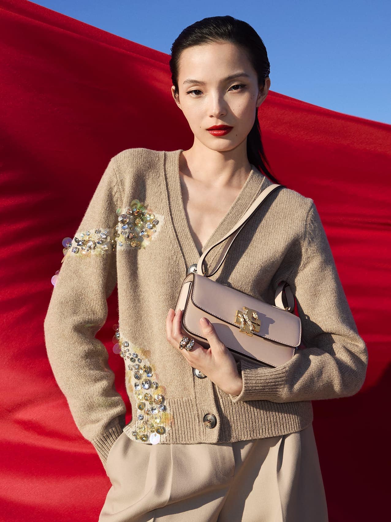 Asian female model wearing a beige embellished cardigan holding a light pink shoulder bag against a bright red fabric backdrop.